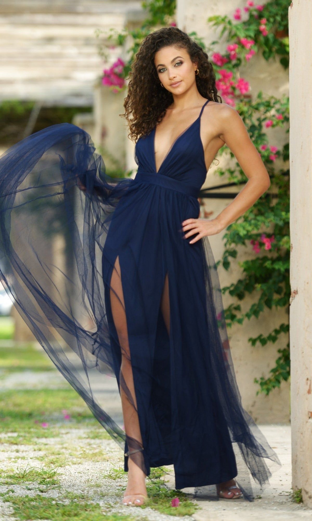 A woman stands outdoors by a wall with green vines and pink flowers, wearing the Velvi Plunging V-Neck Long Tulle Prom Dress Sydney—a sleeveless, deep blue, floor-length gown featuring a sheer overlay.